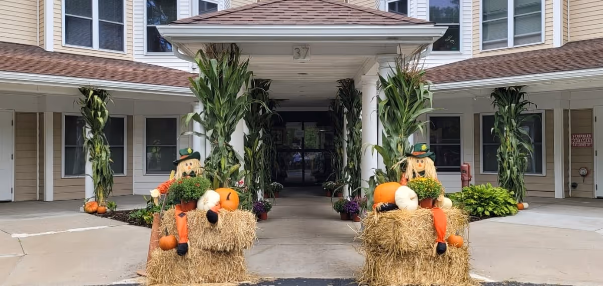 Entrance of Rockridge Retirement Community decorated with cornstalks tied to columns and two scarecrow figures sitting on hay bales holding pumpkins and flowers, with the building facade visible in the background.