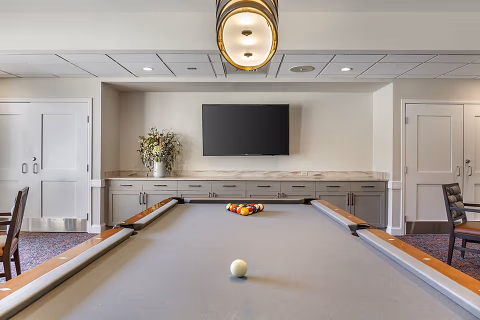 Interior view of a recreational room featuring a pool table with billiard balls arranged for a game. The room has a flat-screen TV mounted on the wall above a long cabinet with drawers and a vase of flowers. There are chairs and tables on either side of the pool table, and the ceiling has recessed lighting and a hanging light fixture.
