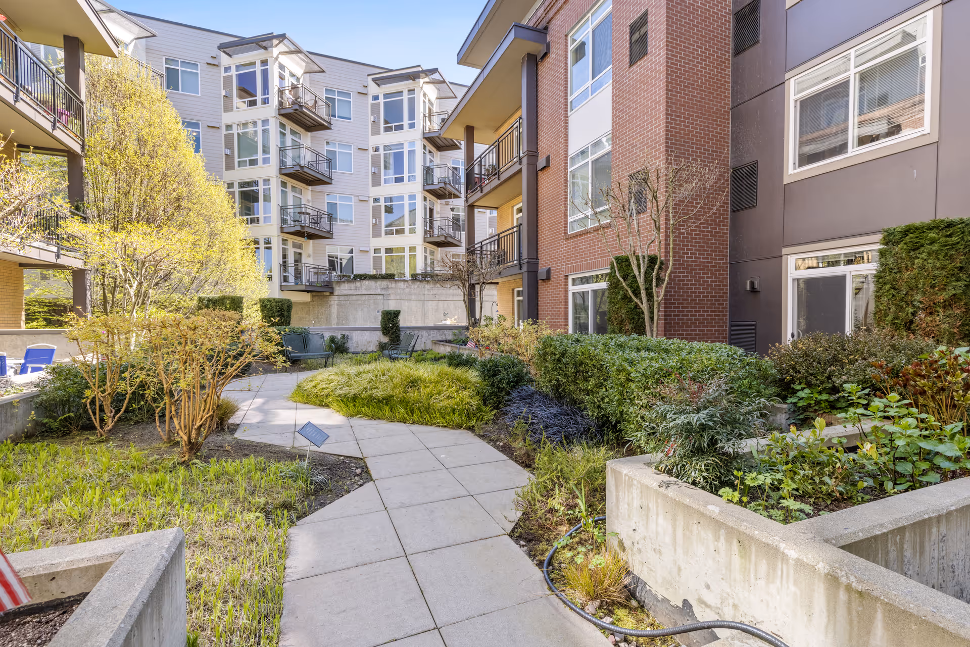 Outdoor courtyard area of a senior living facility with a paved walkway surrounded by landscaped greenery, bushes, and small trees. The courtyard is bordered by multi-story residential buildings with balconies and large windows.