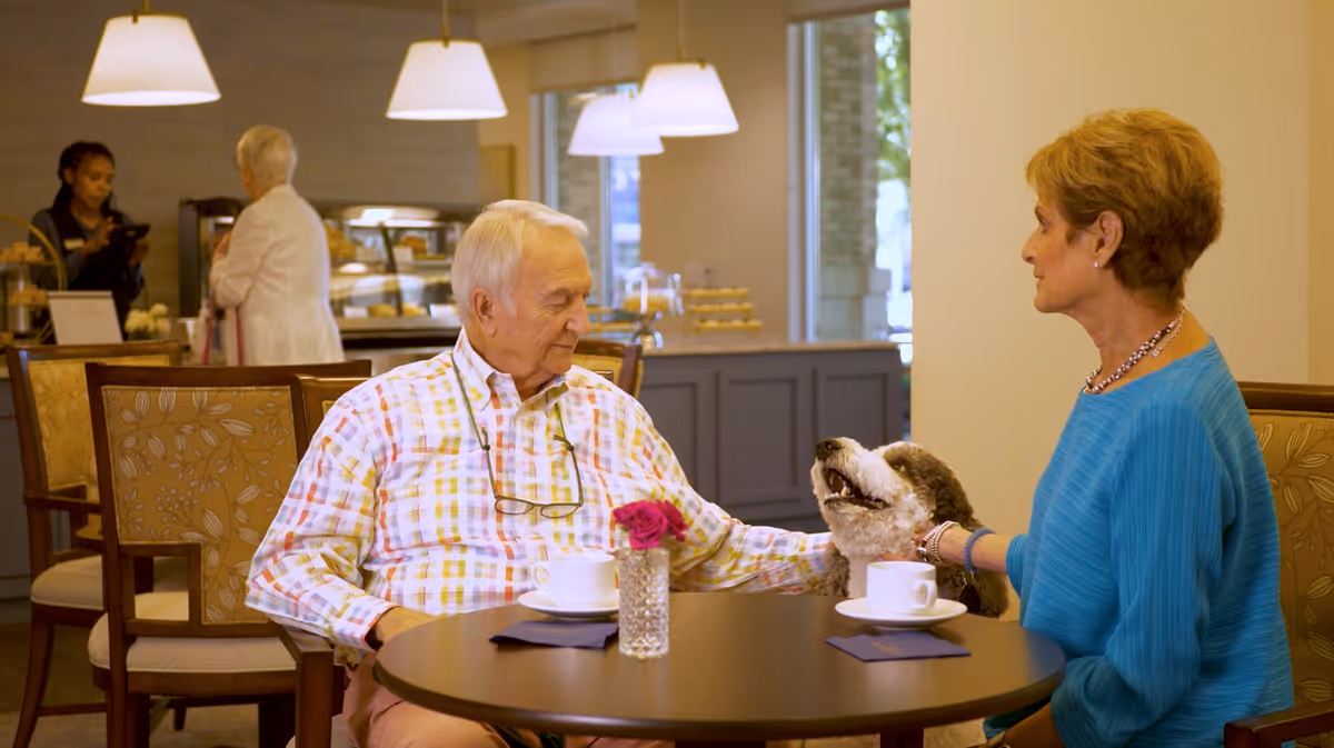 An elderly man and woman sitting at a round table in a cafe or dining area, with two white coffee cups on the table. The man is petting a friendly dog sitting between them. In the background, two women are standing near a counter with baked goods and a coffee machine. The setting is warm and inviting with pendant lights hanging from the ceiling.