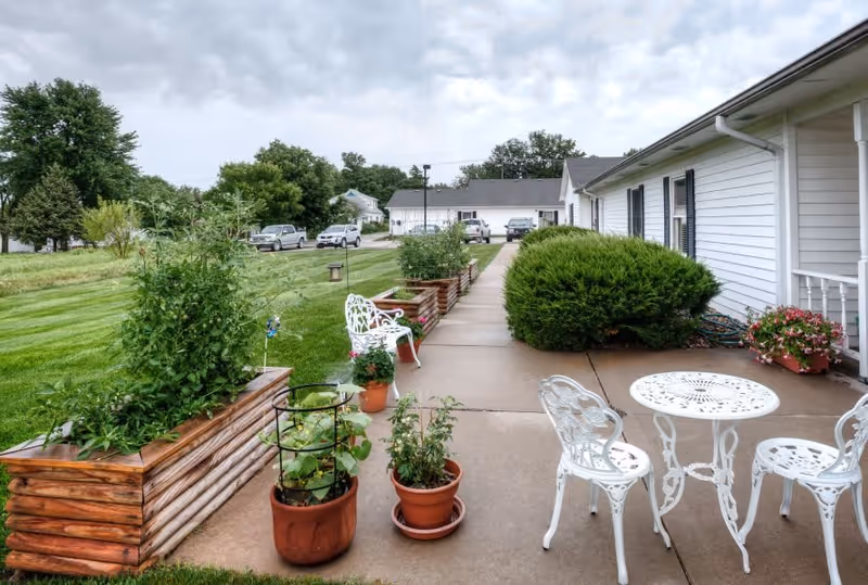 Outdoor patio area at Vintage Park At Louisburg featuring a concrete walkway with white metal table and chairs, potted plants, raised wooden garden beds with greenery, and a white building with black shutters on the right side under a cloudy sky.