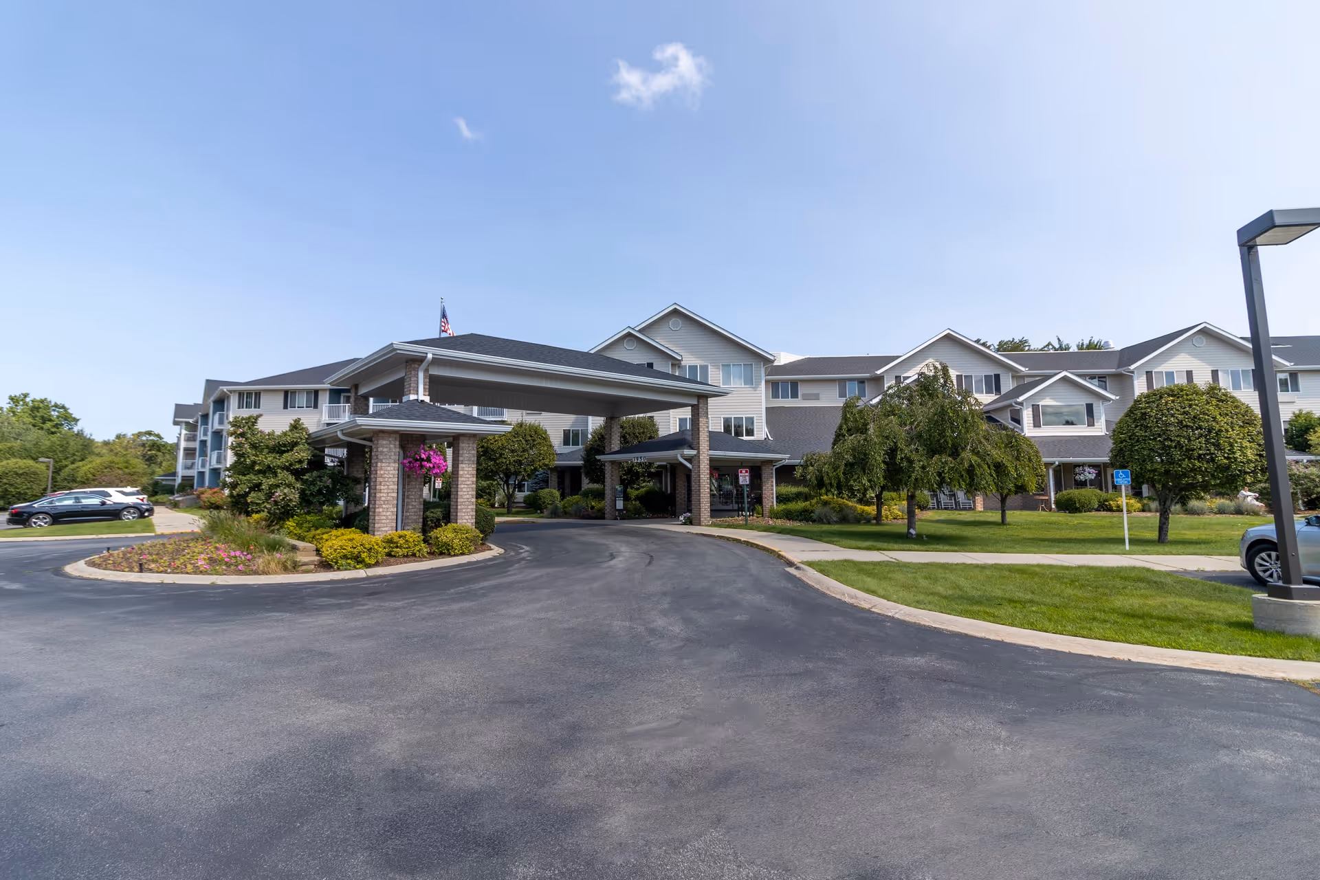 Front exterior view of Independence Village of Traverse City, showing a large building with multiple windows, a covered entrance supported by brick pillars, landscaped greenery, and a clear blue sky.