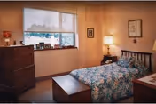 A cozy bedroom with a single bed covered in a floral bedspread, a wooden chest at the foot of the bed, a wooden dresser with framed photos on top, and a window with a white roller shade partially down. The walls are painted a warm beige color and decorated with framed pictures. A bedside table with a lamp is next to the bed.