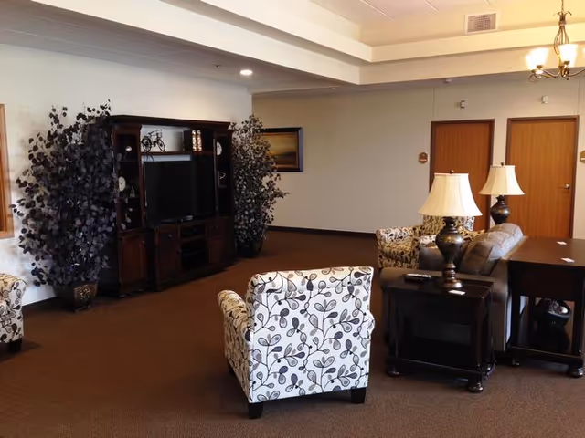 A cozy living room area in a senior living facility with patterned armchairs, a sofa, two table lamps on dark wooden side tables, a large wooden entertainment center with a TV, and two tall decorative plants on either side of the entertainment center. The room has beige walls, brown carpet, and two closed wooden doors in the background.