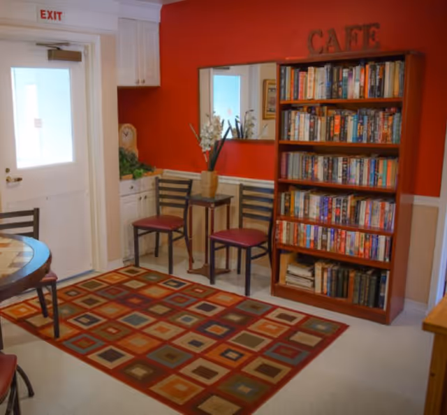Small assisted-living common room with a bookshelf labeled "CAFE", chairs, a patterned rug and an exit door.