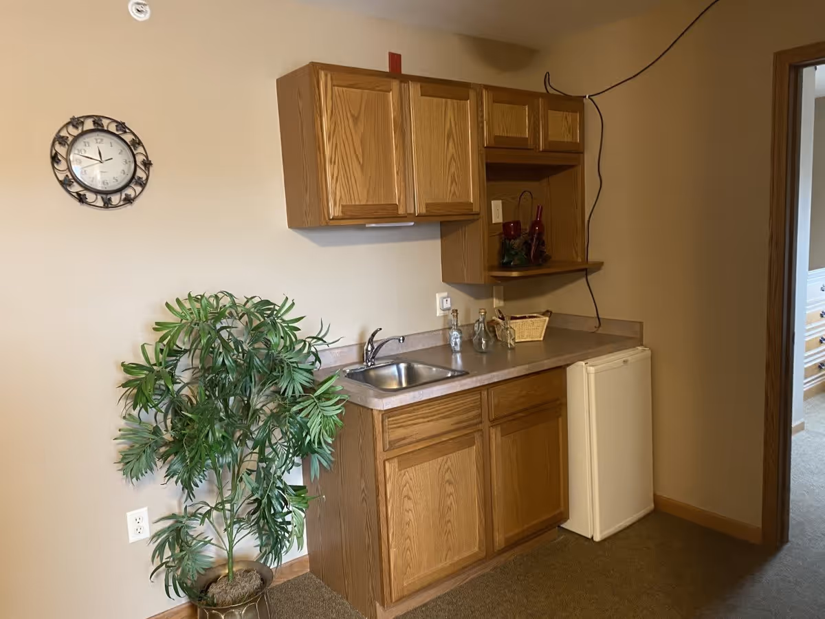 Small kitchenette area with wooden cabinets, a stainless steel sink, a mini refrigerator, a potted green plant, and a wall clock. The countertop has decorative bottles and a small basket. The walls are beige and there is an open doorway leading to another room.