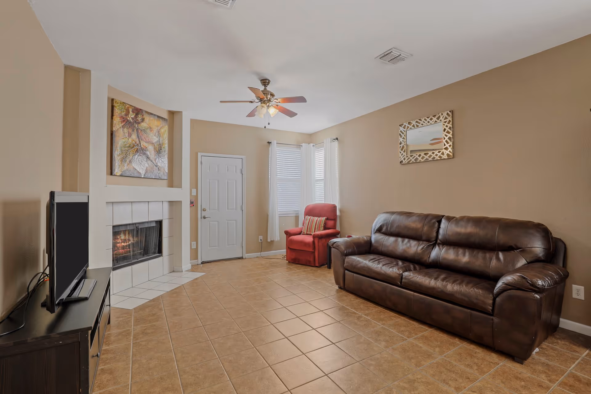 A living room with beige walls and tiled floor featuring a brown leather sofa, a red armchair with a striped pillow, a fireplace with a painting above it, a ceiling fan with lights, a TV on a black stand, and a door with two windows covered by white curtains.