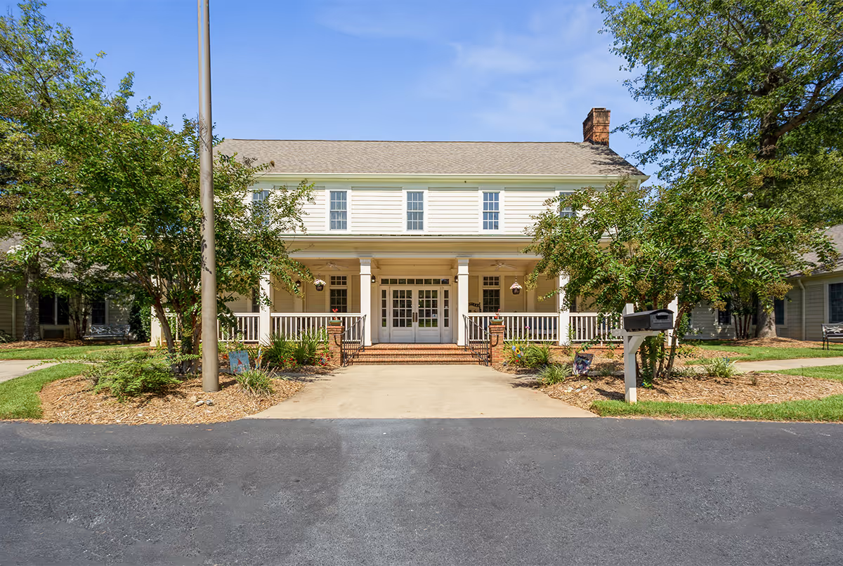 Front exterior view of a two-story building with white siding, a covered porch with white railings and columns, brick steps leading to double glass doors, surrounded by trees and landscaping under a clear blue sky.