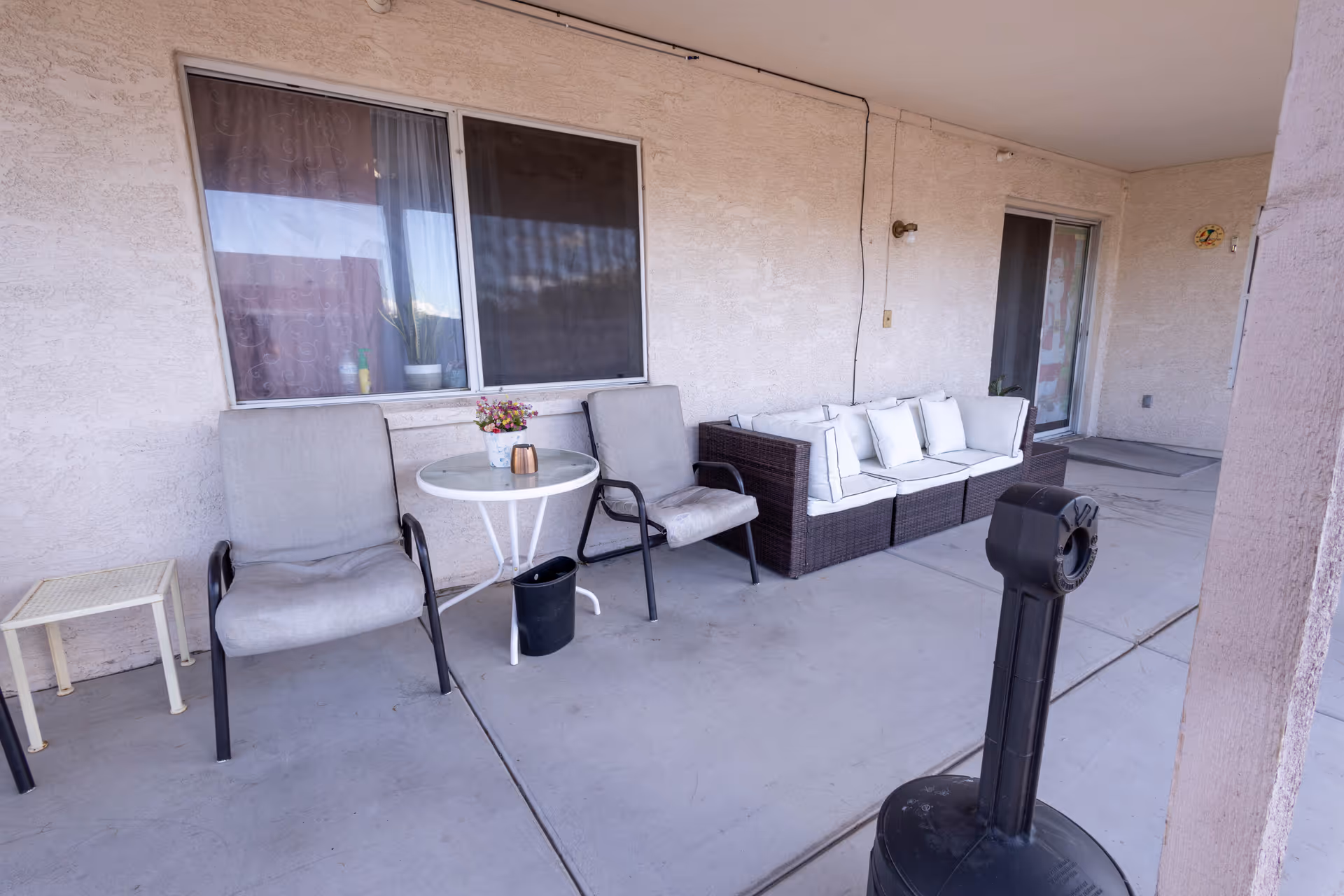 Covered outdoor patio with two chairs, a small round table, and a cushioned wicker sofa in front of windows and a sliding door.