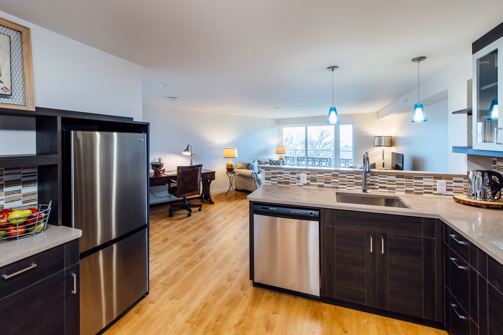 Modern kitchen area with dark wood cabinets, a stainless steel refrigerator, dishwasher, and sink. The kitchen opens into a living space with a desk, chair, sofa, lamps, and large windows letting in natural light.