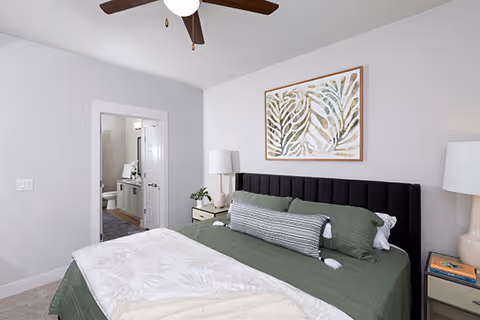 A bedroom with a large bed featuring a black upholstered headboard, green bedding, and a long decorative pillow. There are two bedside tables with lamps and a framed botanical artwork on the wall above the bed. An open door reveals a bathroom with a vanity and toilet.