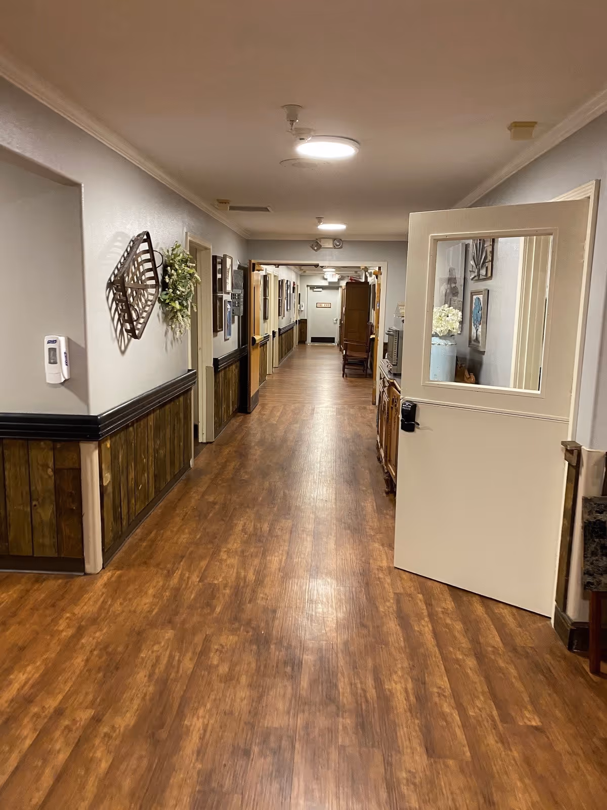 A long indoor hallway with wooden flooring and wood-paneled lower walls. The hallway has several doors on both sides, some open, and is decorated with wall art and a small wreath. Ceiling lights illuminate the corridor, and there is a hand sanitizer dispenser mounted on the wall.