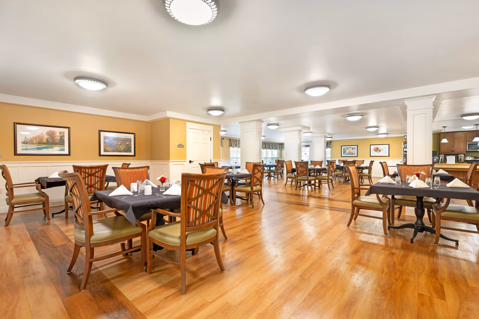 Spacious dining room with multiple set tables and wooden chairs on a polished hardwood floor and framed artwork on the walls.