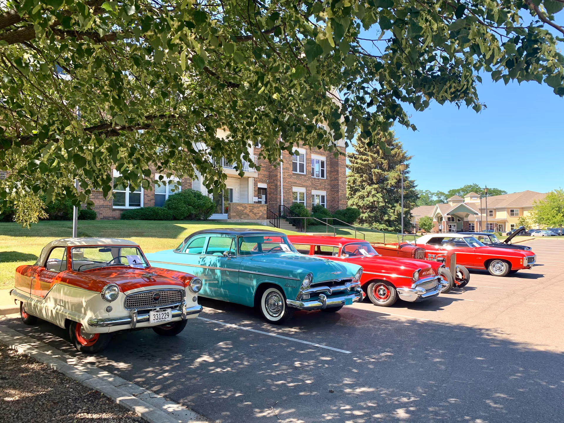 Several colorful vintage cars parked under trees in front of a brick building on a sunny day.