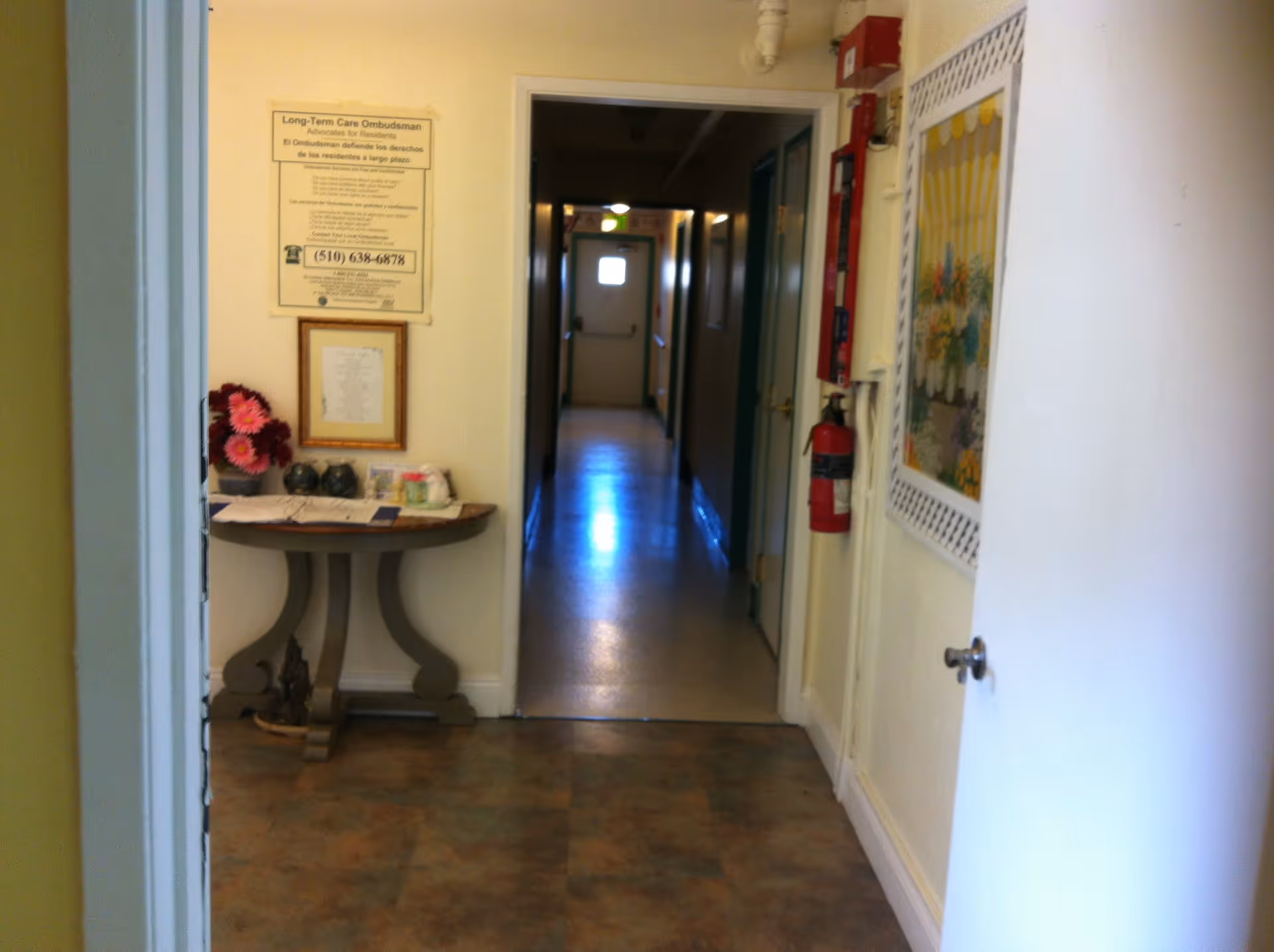 View of a hallway in a senior living facility with a small table on the left holding flowers and other items. A sign about Long-Term Care Ombudsman is mounted on the wall above the table. The hallway has several doors along it and a fire extinguisher is mounted on the right wall. A framed floral picture hangs on the right wall near an open door.