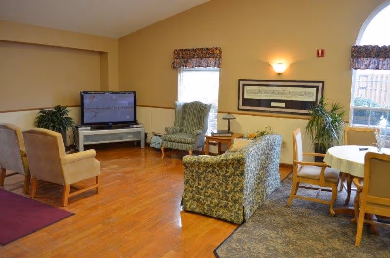 A cozy living room area in a senior living facility with wooden flooring, a patterned sofa, two beige armchairs, a green striped armchair, a TV on a stand, a small side table with a lamp, a round dining table with chairs, and large windows with floral valances letting in natural light.