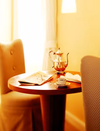 Small round wooden table by a window with two upholstered chairs, a silver teapot, a cup and saucer, and a folded newspaper.