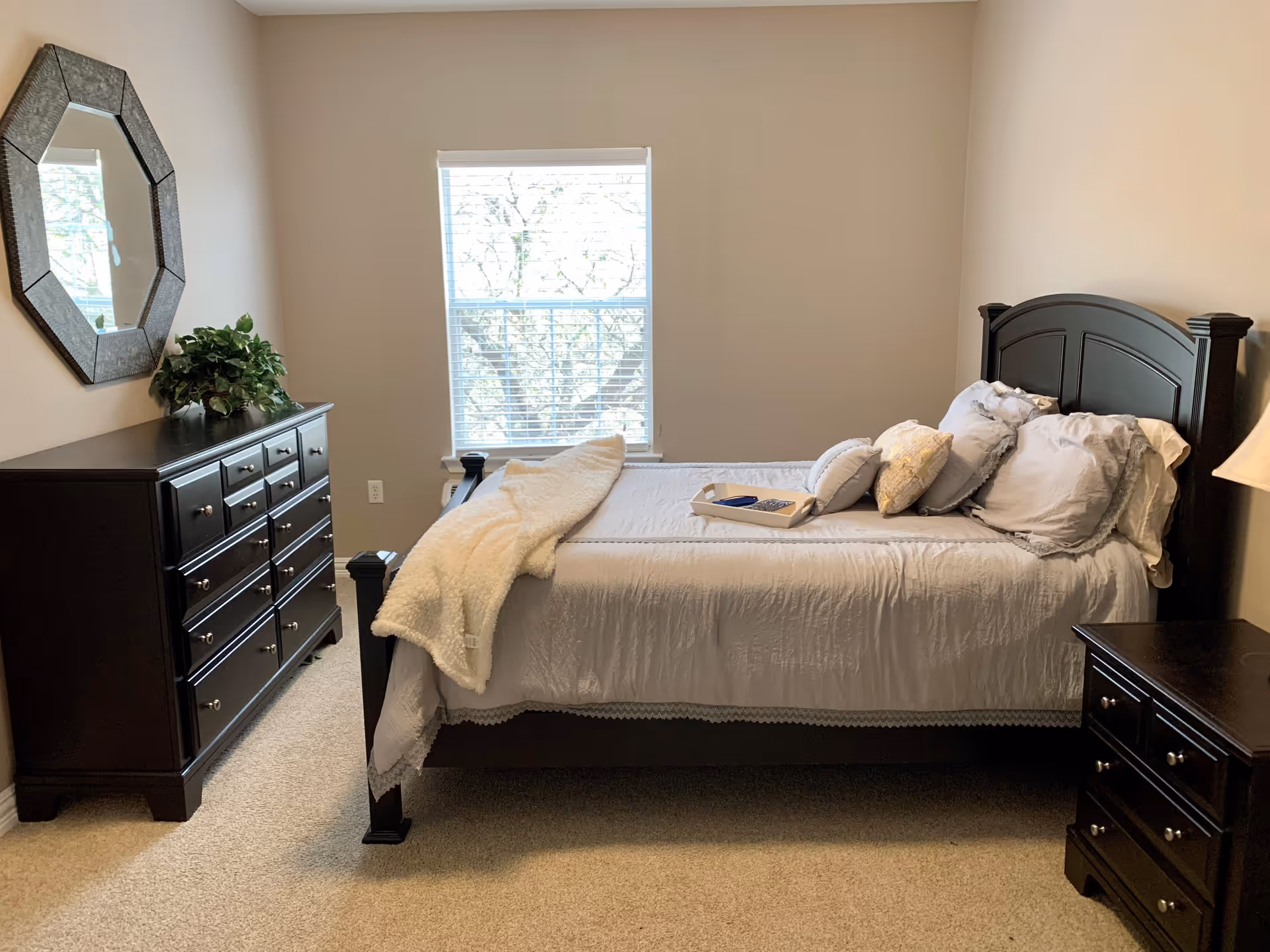 A neatly made bed with white bedding and multiple pillows in a bedroom. There is a dark wooden dresser with a decorative mirror and a plant on top to the left of the bed. A window with blinds is centered on the wall behind the bed, and a dark wooden nightstand with a lamp is on the right side of the bed. The room has beige walls and carpeted flooring.