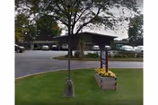 Grassy lawn with a small tree and flower planter in front of a single-story mid-century building and circular driveway with parked cars.