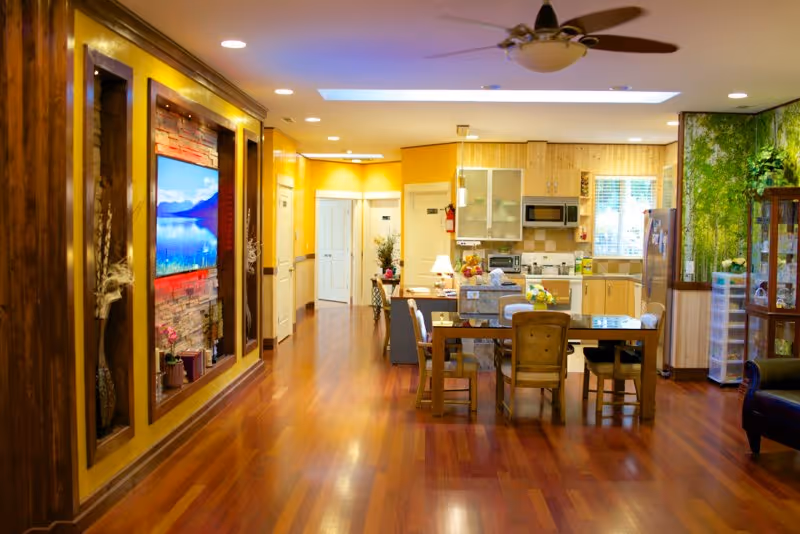 A bright and spacious interior room featuring a wooden floor and a dining table with chairs in the center. The room includes a kitchen area with cabinets, a microwave, and a refrigerator. On the left wall, there is a mounted flat-screen TV surrounded by decorative wooden panels and stone accents. The right side has a green nature-themed wall mural and a glass cabinet. The room is well-lit with ceiling lights and a ceiling fan.