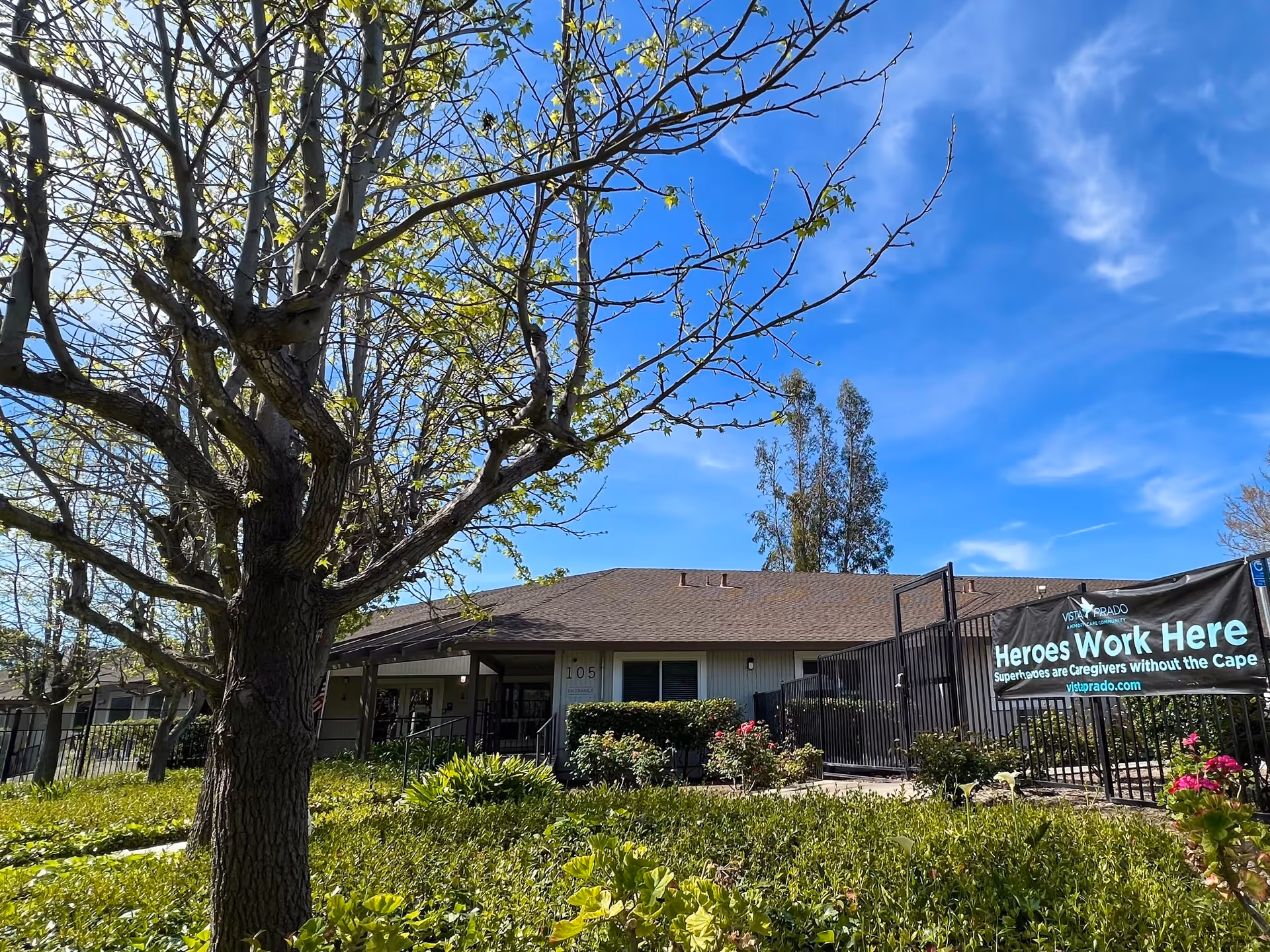 Front exterior of a single-story senior living building with landscaped grounds, a large tree in the foreground, and a 'Heroes Work Here' banner on the fence.