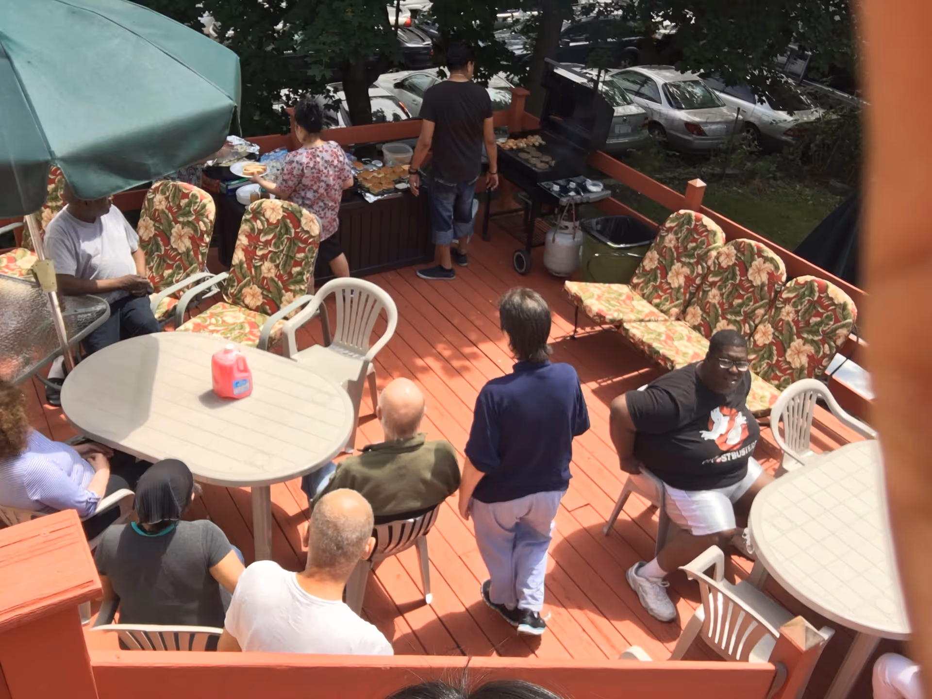 A group of people gathered on a wooden outdoor deck with red flooring. Some are seated around plastic tables and chairs, while others are standing near a grill where food is being prepared. The deck has cushioned floral-patterned chairs and is surrounded by trees and parked cars in the background.