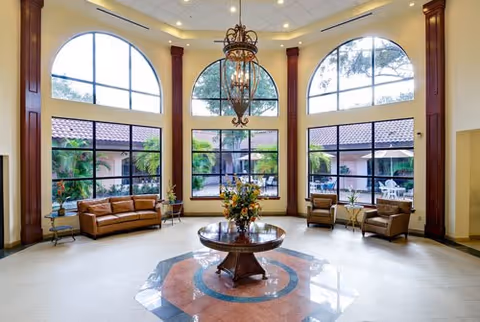 Spacious senior living facility lobby with large arched windows, a central round table with flowers, chandelier, sofas and armchairs.