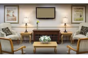 A symmetrical lounge with upholstered chairs, a wall-mounted TV, side tables with lamps, and a coffee table holding a floral arrangement.