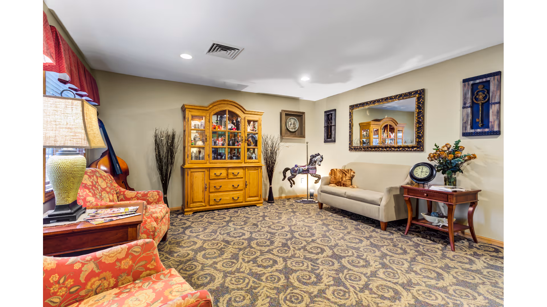 A cozy living room with patterned carpet, a beige sofa with a brown pillow, two red floral armchairs, a wooden side table with a lamp and magazines, a wooden cabinet filled with decorative items, a large ornate mirror on the wall, and wall art including a framed key and a circular picture.