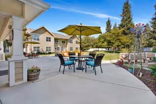 Outdoor patio area at Dale Commons featuring a round table with four chairs and a large green umbrella. The patio is surrounded by a garden with raised planting beds and a decorative windmill. In the background, there is a two-story building with beige siding and multiple windows, set against a clear blue sky with some trees.