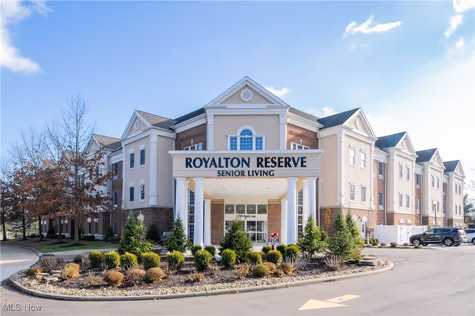 Front exterior view of the Royalton Reserve Senior Living facility, a large three-story building with a combination of beige and brick facade, white columns at the entrance, and landscaped bushes and trees in front under a clear blue sky.