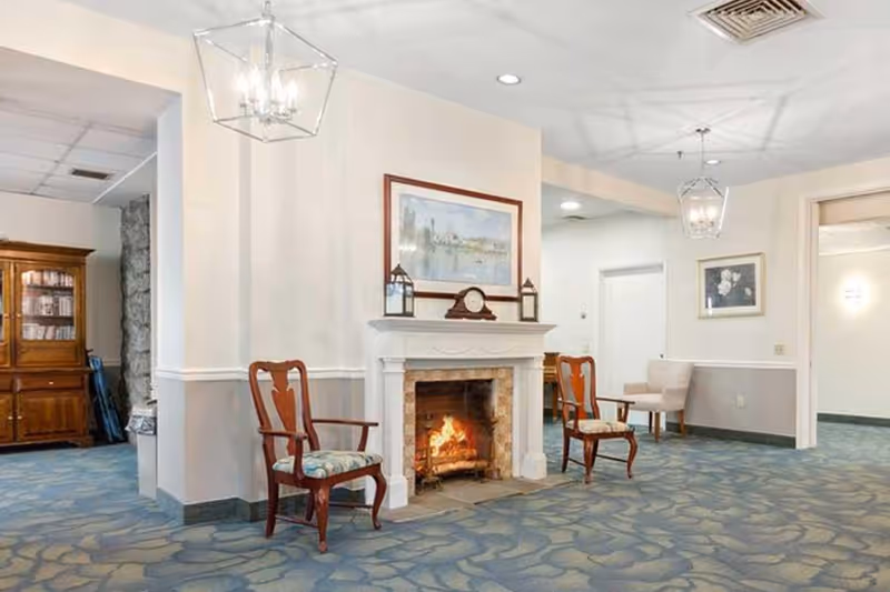 A cozy senior living facility common area featuring a lit fireplace with a mantel decorated with a clock and two lanterns. Two wooden armchairs with patterned cushions are placed on either side of the fireplace. The room has patterned blue carpet, white walls with a chair rail, and framed artwork above the fireplace and on the adjacent wall. There is a wooden cabinet with glass doors in the background and a beige armchair with a small table near a doorway.