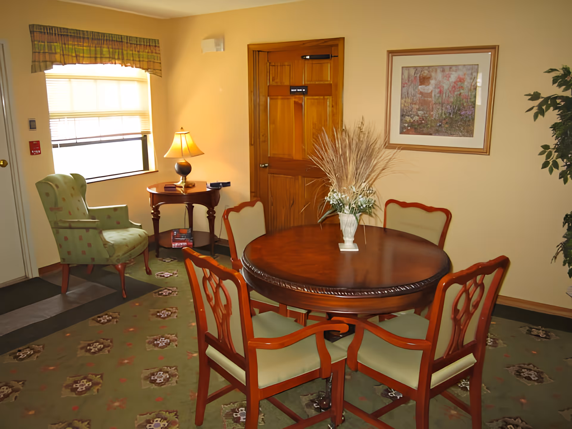 A cozy room with a round wooden table surrounded by four chairs with green cushions. On the table is a vase with dried decorative grass. To the left, there is a green upholstered armchair next to a small wooden side table with a lamp. The room has a window with blinds and a valance, a wooden door, a framed floral painting on the wall, and a patterned green carpet.