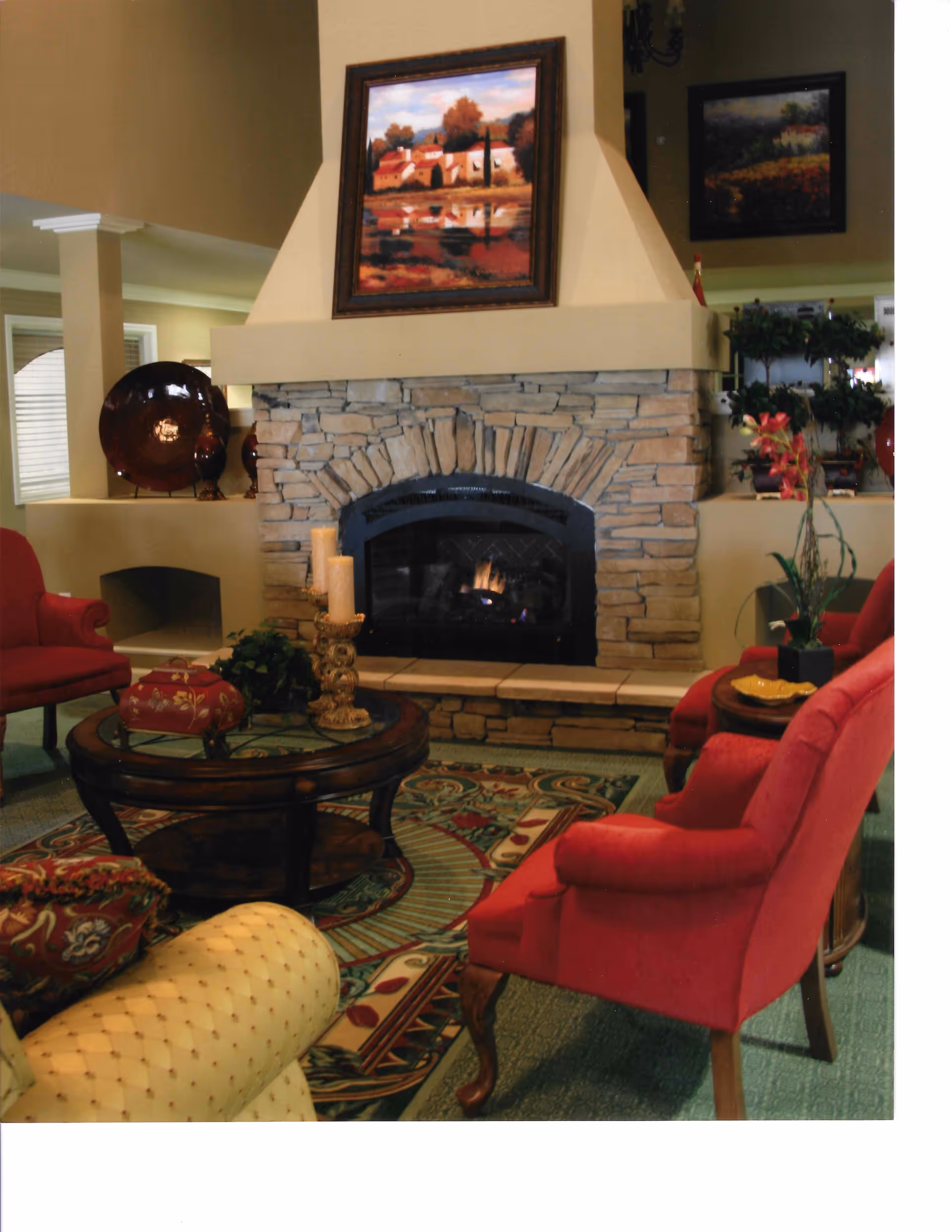 Cozy living room area with a stone fireplace in the center, a painting above the fireplace, red upholstered chairs, a round wooden coffee table with candles and decorative items, and a patterned rug on the floor.