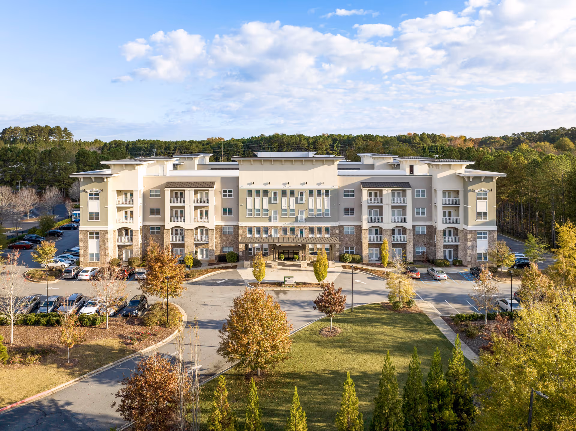 Front exterior of a multi-story senior living building with parking and landscaped grounds under a partly cloudy sky.