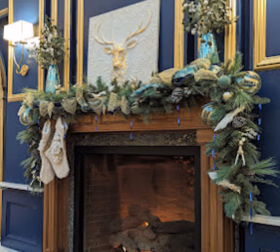 A decorated fireplace mantel with evergreen garland, ornaments, hanging stockings, and a deer relief on a blue-paneled wall.