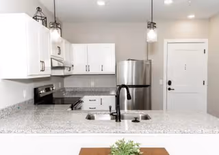 Modern kitchen with white cabinets, stainless steel refrigerator and stove, granite countertops, and a double sink with a black faucet. Pendant lights hang from the ceiling and a white door is visible in the background.