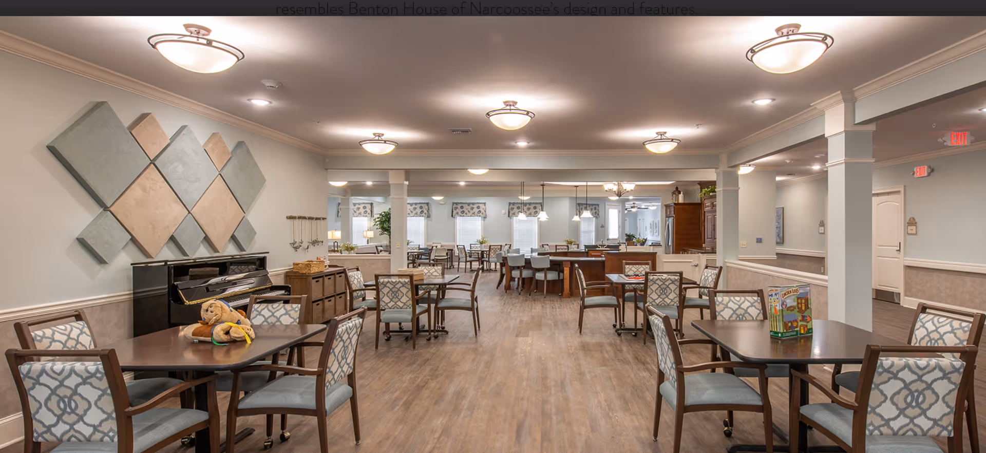 A spacious and well-lit common dining area in a senior living facility with multiple tables and chairs arranged neatly. The room features a piano against one wall, decorative wall art, and soft overhead lighting. The flooring is wood-style, and the walls are painted in light colors with crown molding. There are windows with patterned valances and a kitchen area visible in the background.