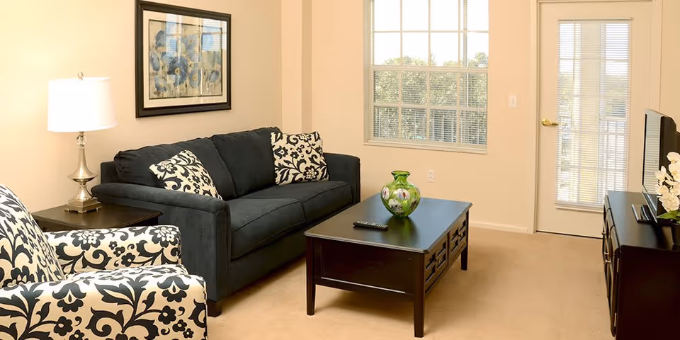 A cozy living room with a dark blue sofa and two patterned cushions, a matching patterned armchair, a dark wooden coffee table with a green decorative vase and a remote control on it, a side table with a lamp, a framed floral artwork on the wall, a window with blinds, and a door with glass panels leading outside.