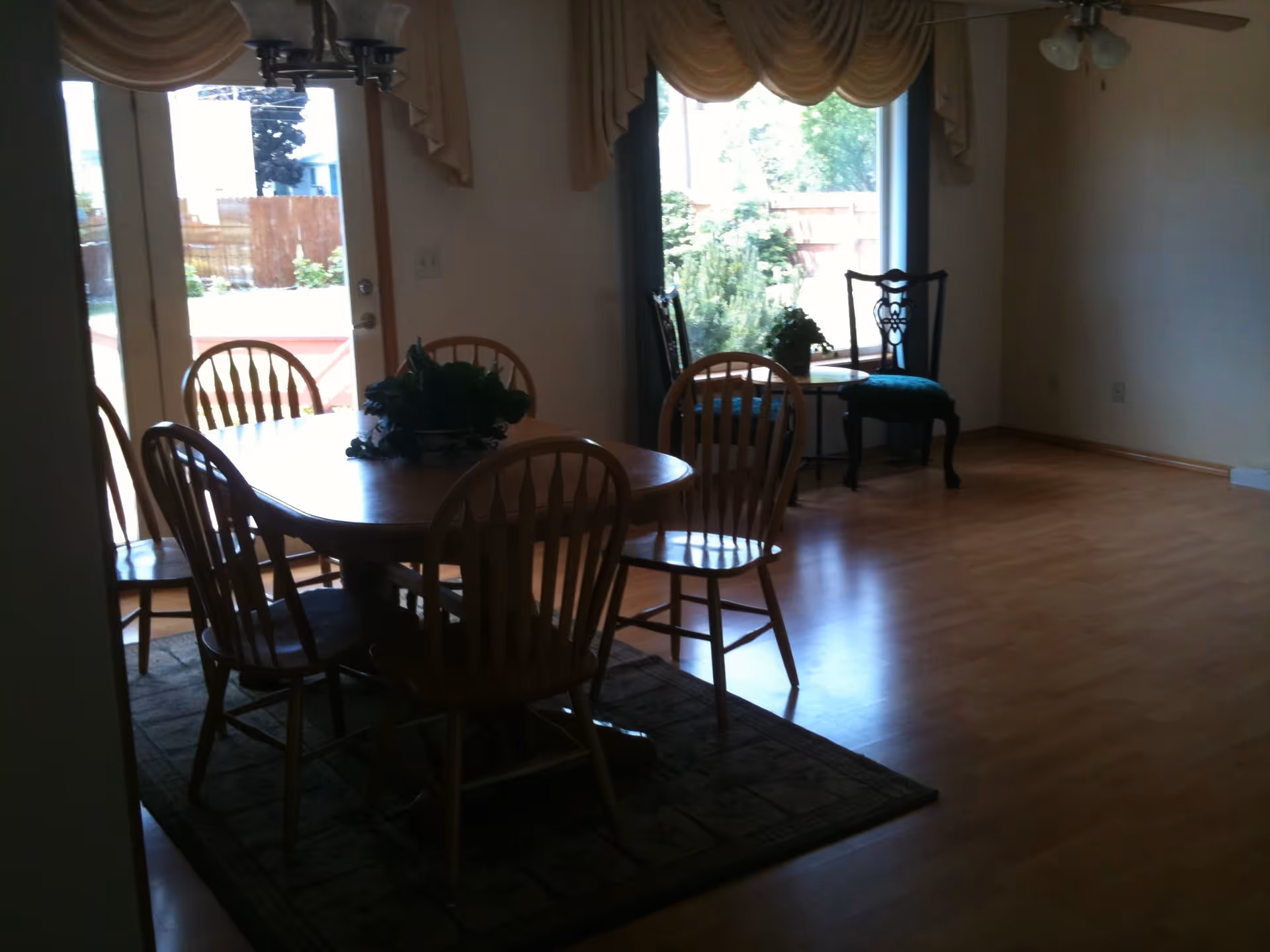 Dining area with a wooden table and six matching wooden chairs on a rug. There is a glass door and a large window with beige curtains, letting in natural light. A small table with two decorative chairs is placed near the window.
