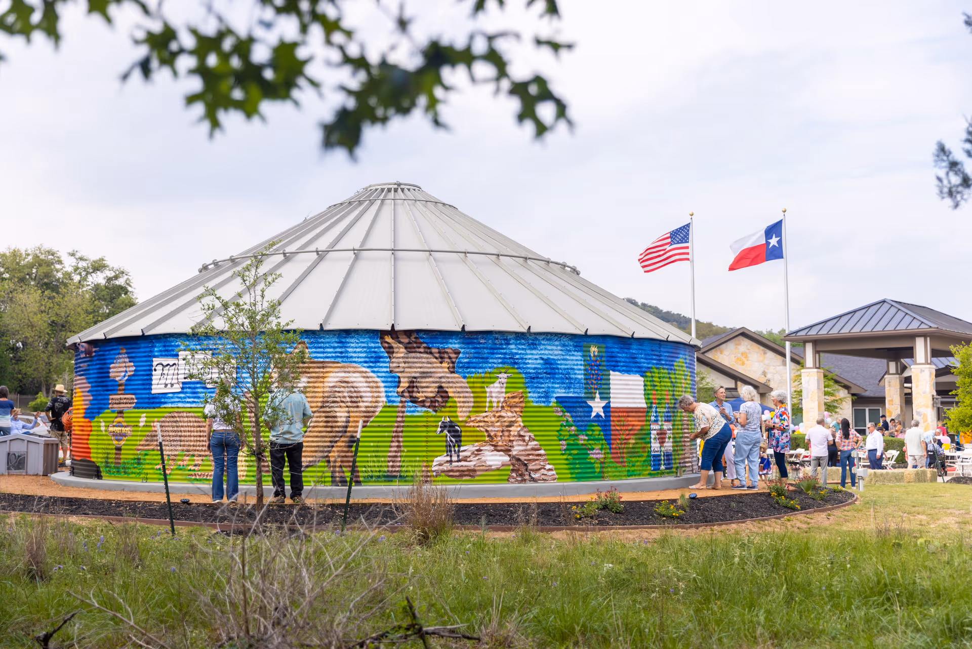 Outdoor scene at Alexis Pointe of Wimberley Senior Living showing a round metal building with a colorful mural of animals and the Texas flag. Several people are gathered around the building and near a pavilion with two flags, the American flag and the Texas state flag, flying on flagpoles.