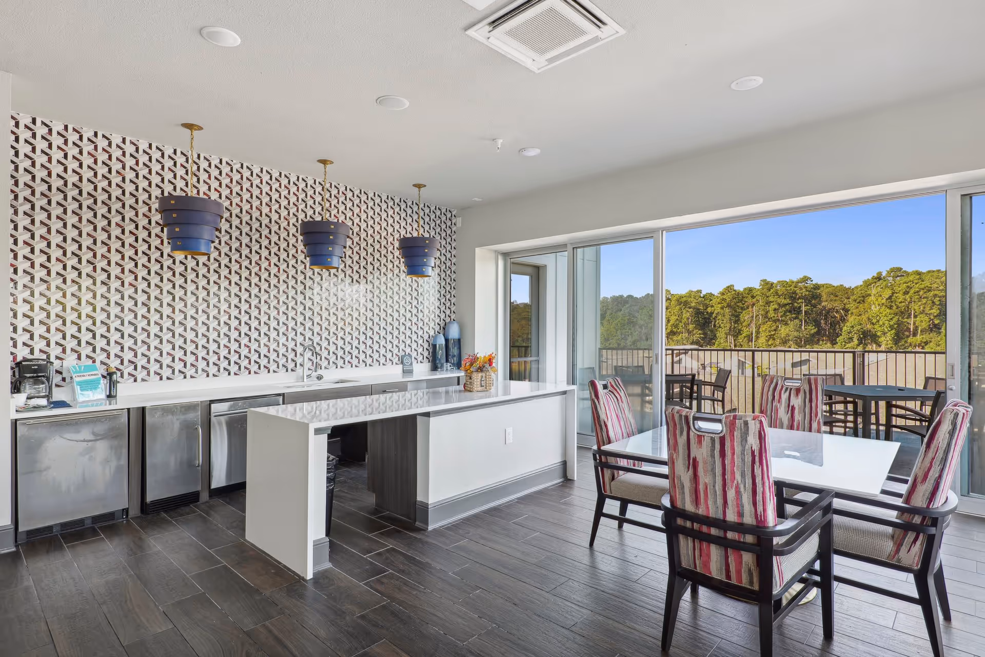 Modern common area with a kitchenette and island, pendant lights, and a dining table by floor-to-ceiling windows overlooking trees.