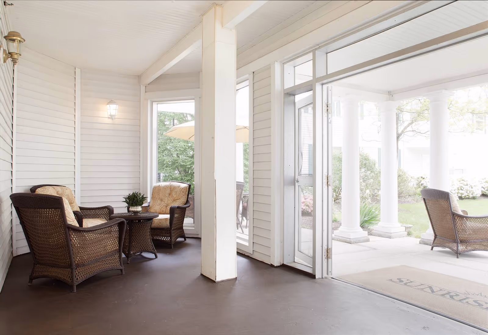 A covered outdoor seating area with wicker chairs and cushioned seats arranged around a small table. The area is adjacent to a garden with white columns and greenery visible outside. The space has white paneled walls and a glass door leading outside.