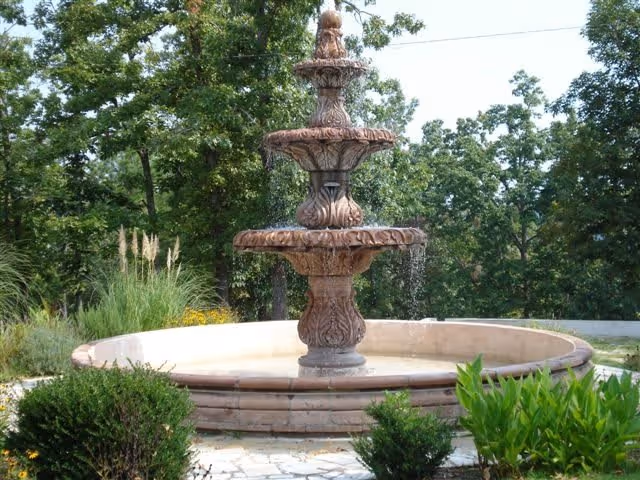 Three-tiered ornamental stone fountain in a landscaped outdoor area with surrounding shrubs and trees.