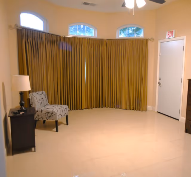 A small room with beige walls and tiled floor featuring a single patterned armchair next to a dark wooden side table with a lamp. There are three arched windows covered by long mustard-colored curtains, and a white door with an exit sign above it. A ceiling fan with lights is partially visible at the top.