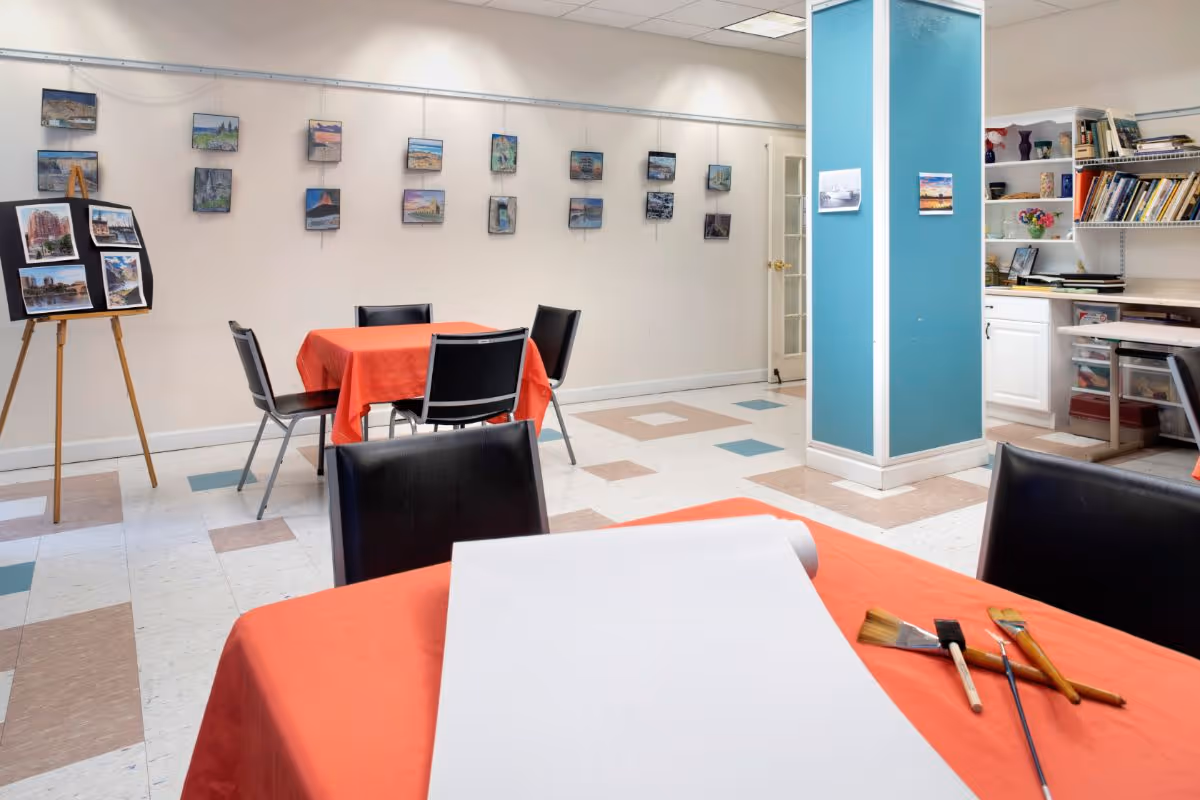 A bright art room with tables covered in orange tablecloths, black chairs, and art supplies including paintbrushes and a blank canvas. The walls display various small paintings and there is an easel with more artwork. A blue pillar stands near shelves filled with books and art materials.