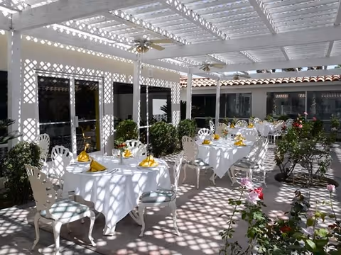 Outdoor dining area with white tables and chairs under a white lattice pergola. Tables are set with white tablecloths, yellow napkins, and glassware. There are plants and flowers around the seating area, and ceiling fans are mounted on the pergola.