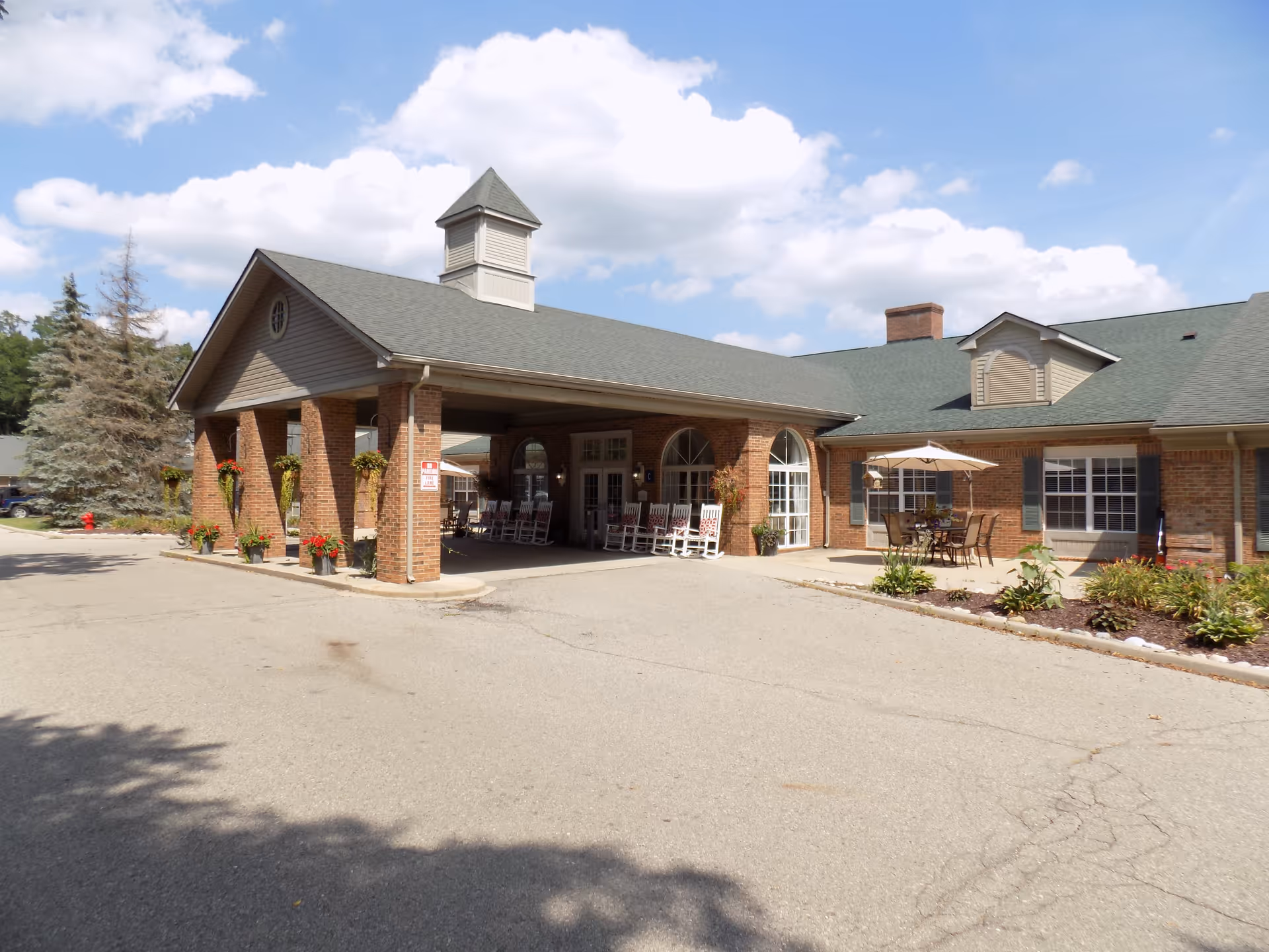 Exterior view of a senior living facility with a covered entrance supported by brick pillars. There are several white rocking chairs under the covered area and a patio with a table, chairs, and an umbrella to the right. The building has a green roof and brick walls, with some landscaping and trees around the driveway.