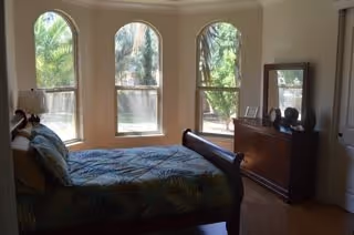 Sunlit bedroom with a bed, dresser, mirror and three arched windows overlooking palm trees.