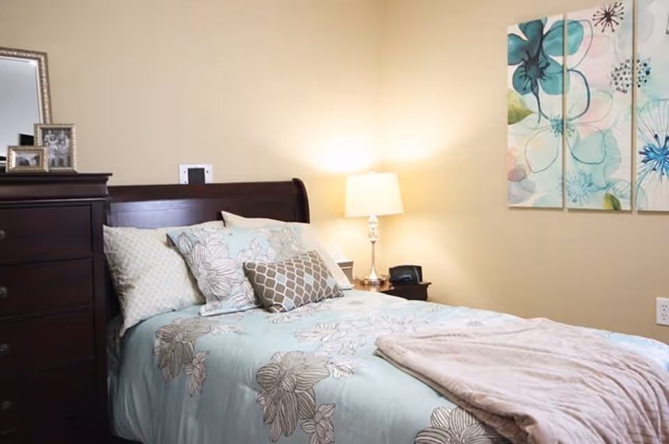 A cozy bedroom with a wooden bed frame and a bed made with light blue floral bedding and multiple pillows. There is a dark wooden dresser with framed photos on top, a bedside table with a lamp and a clock, and a three-panel floral artwork on the beige wall.